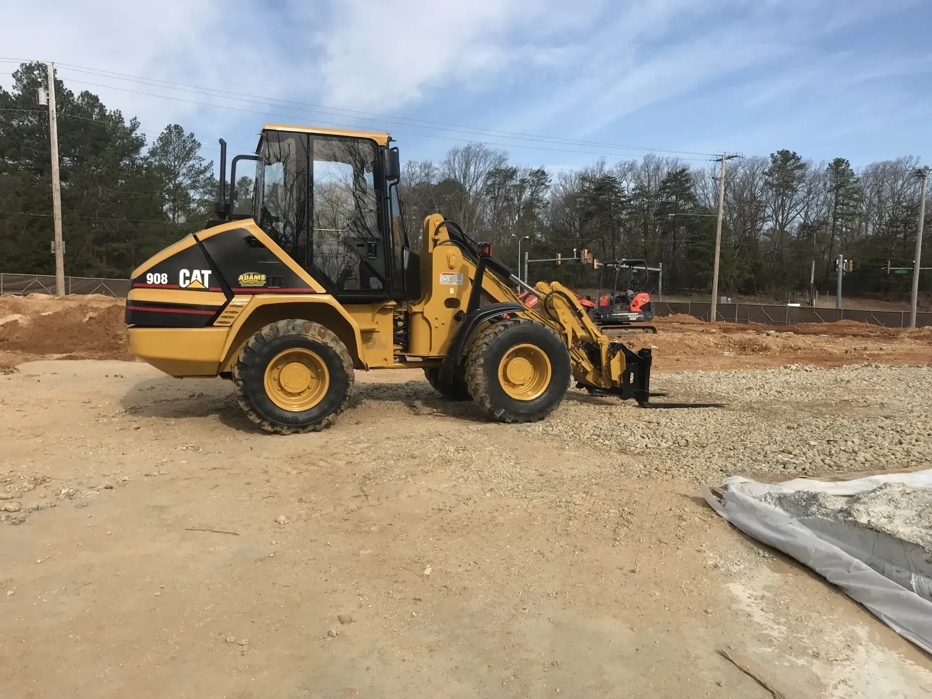 A yellow Caterpillar wheel loader with forks sits on a gravel construction site under a blue sky.