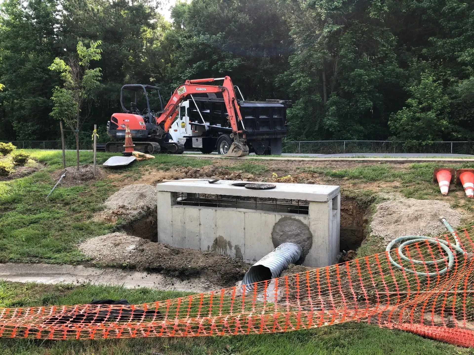 An orange excavator works at a construction site featuring an exposed concrete drainage structure and a metal pipe.