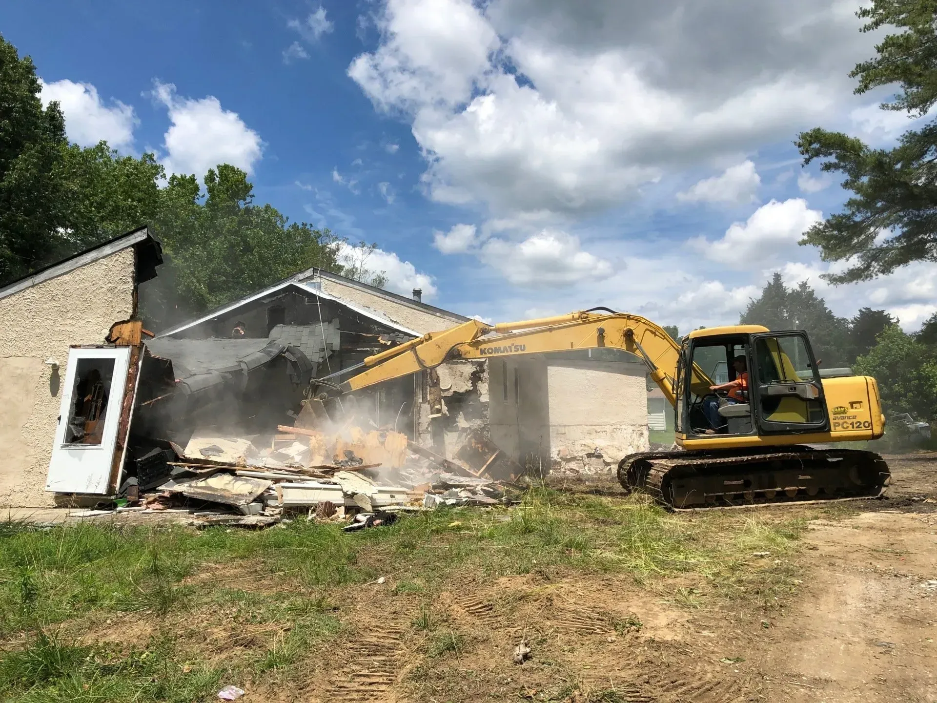 A yellow excavator demolishes a light-colored building under a sunny, cloudy sky.