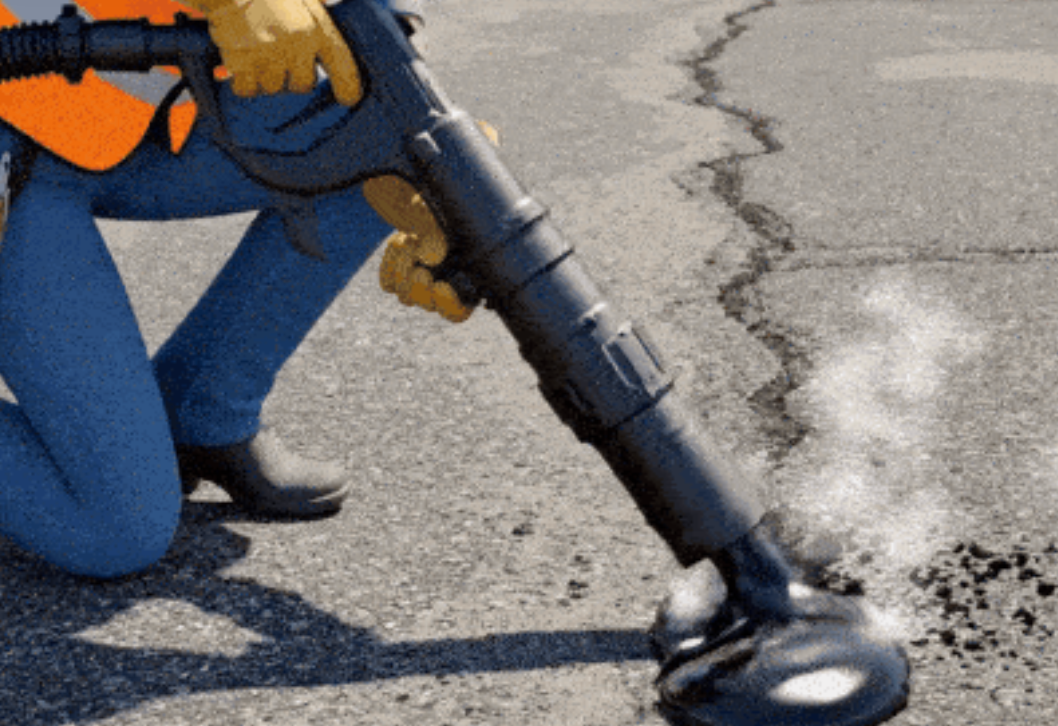 A construction worker in a high-visibility vest uses a hot air lance to clean and dry a crack in an asphalt road.