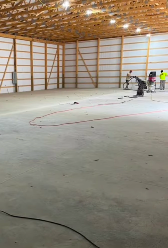 Two workers use industrial floor grinding equipment inside a large, unfinished warehouse with exposed wooden trusses.