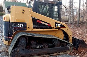 A yellow Caterpillar 277 compact track loader parked on a gravel surface with woods in the background.