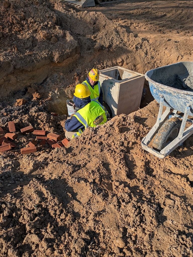 Two workers in high-visibility vests and hard hats install a concrete drainage structure in an excavated trench.