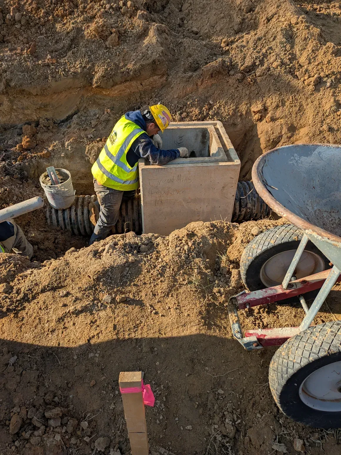 A worker in a high-visibility vest inspects a concrete utility box installed in a dirt trench next to a wheelbarrow.
