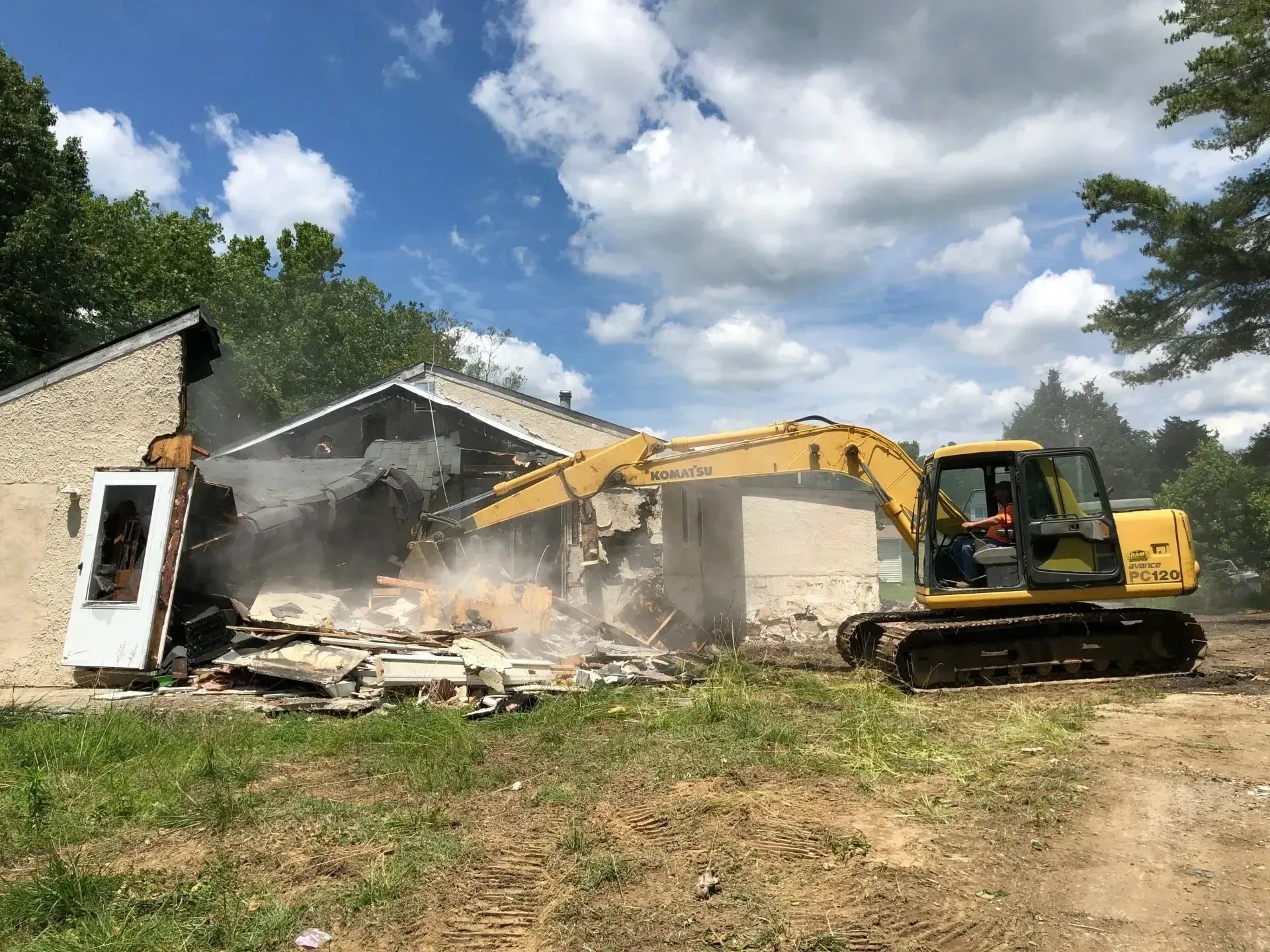 A yellow excavator demolishes a light-colored building, creating a cloud of dust under a blue sky with white clouds.