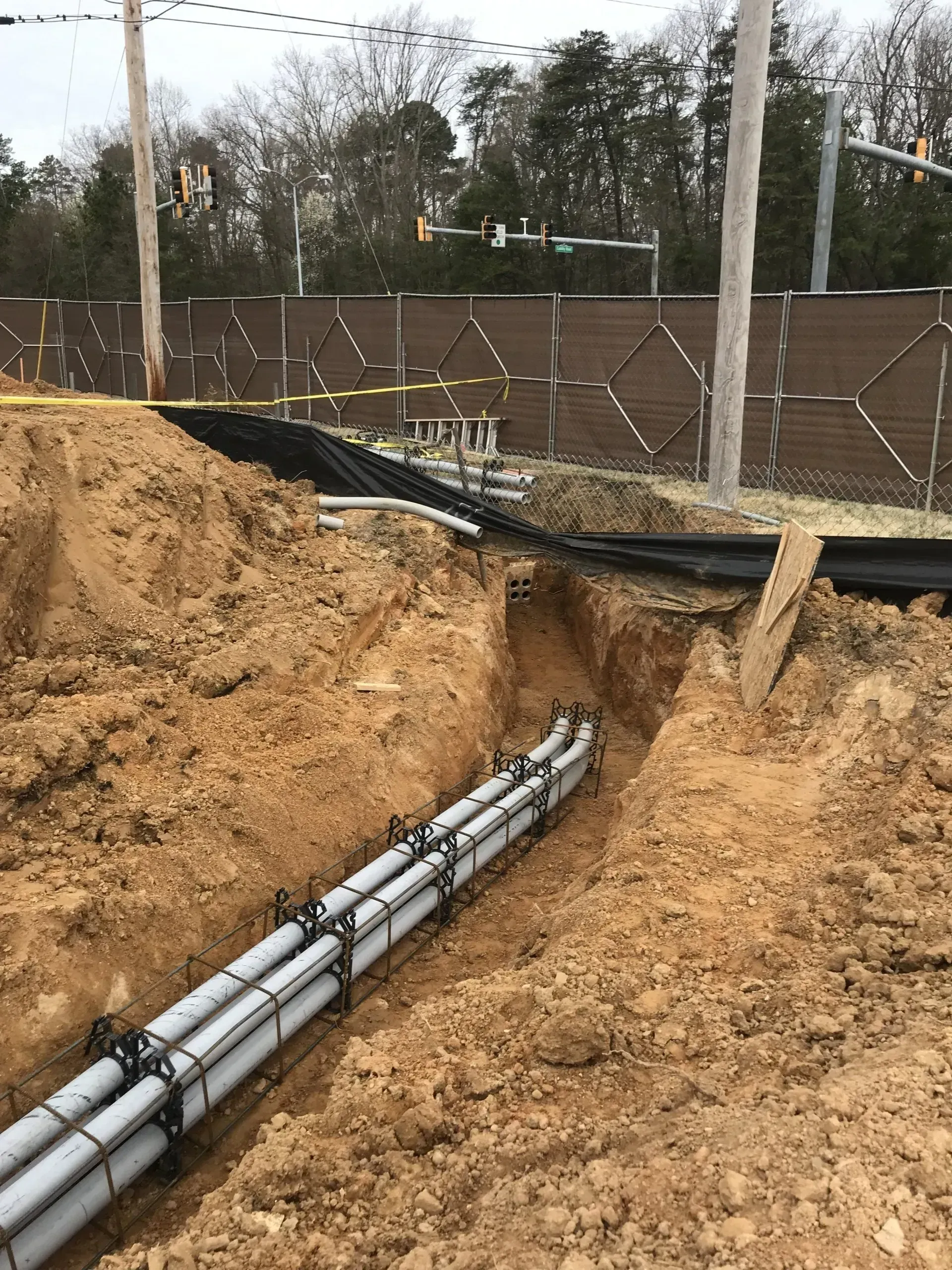 A narrow trench in the ground containing several grey electrical conduits held together by black spacers.