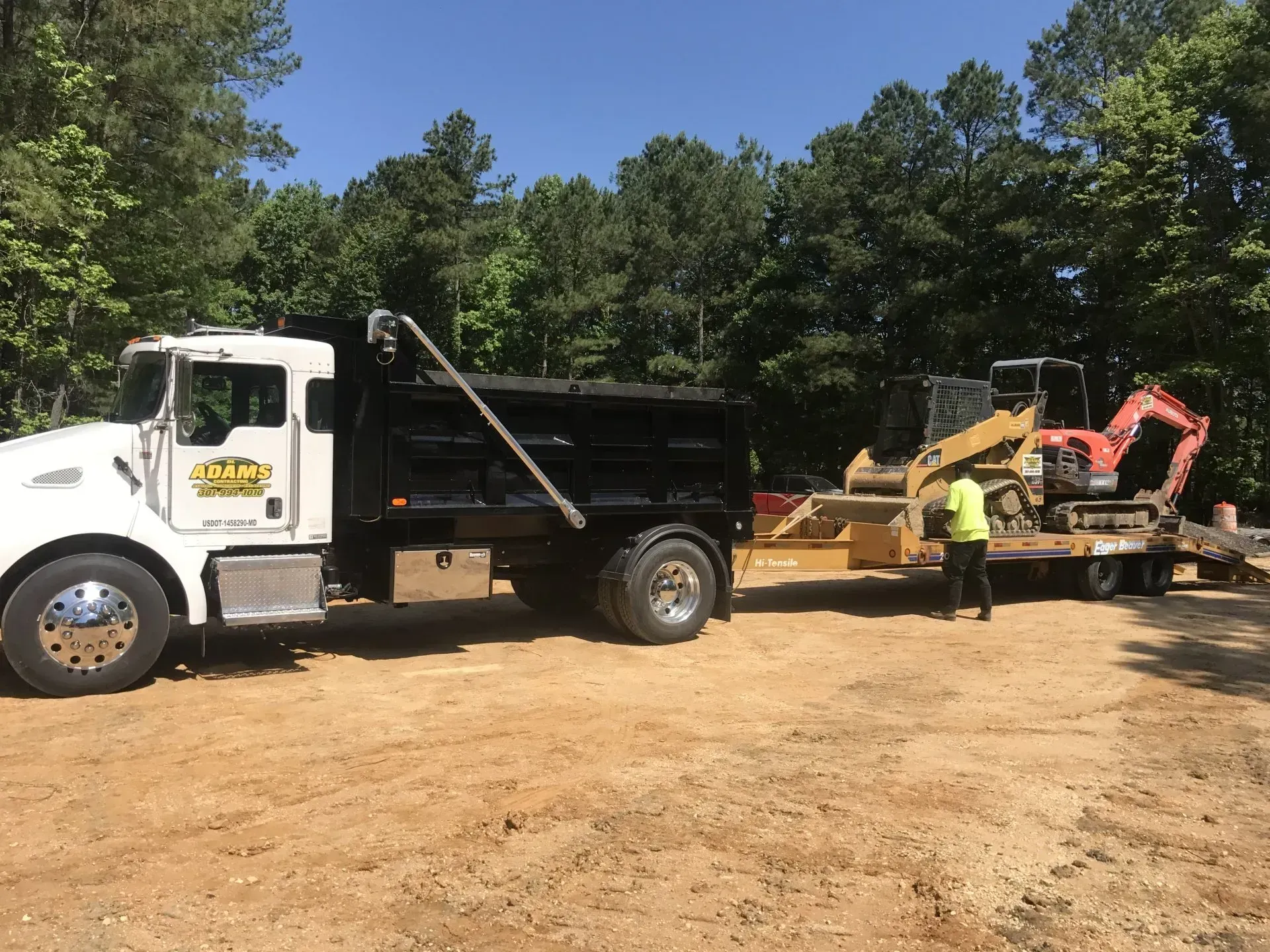 A white dump truck pulls a trailer with a yellow and red excavator on a dirt lot lined with trees.