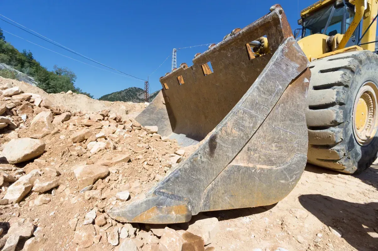 A yellow construction loader bucket rests on a pile of dirt and rocks under a clear blue sky.