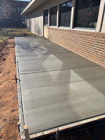 A freshly poured concrete sidewalk adjacent to a brick building wall with large windows, surrounded by dirt.