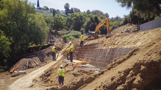Construction workers in high-visibility gear work on a hillside retaining wall with an excavator in a dirt environment.