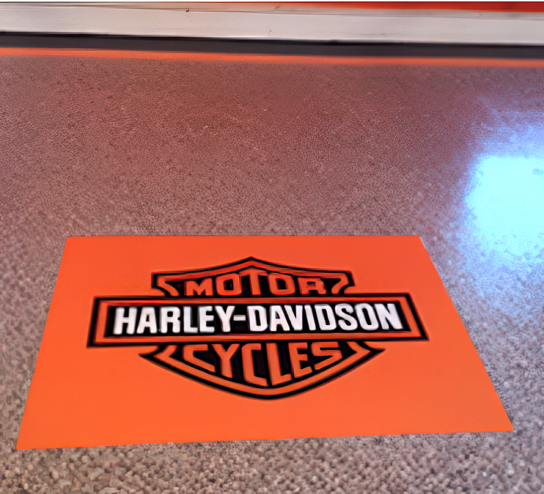 An orange floor mat featuring the black Harley-Davidson logo, placed on a grey speckled floor.