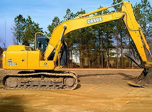 A yellow John Deere excavator sits on a dirt construction site against a backdrop of trees under a blue sky.
