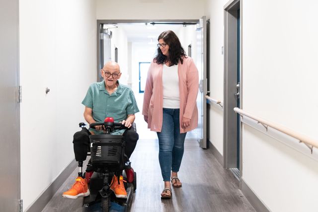 A woman is walking with an elderly man in a mobility scooter down a hallway.