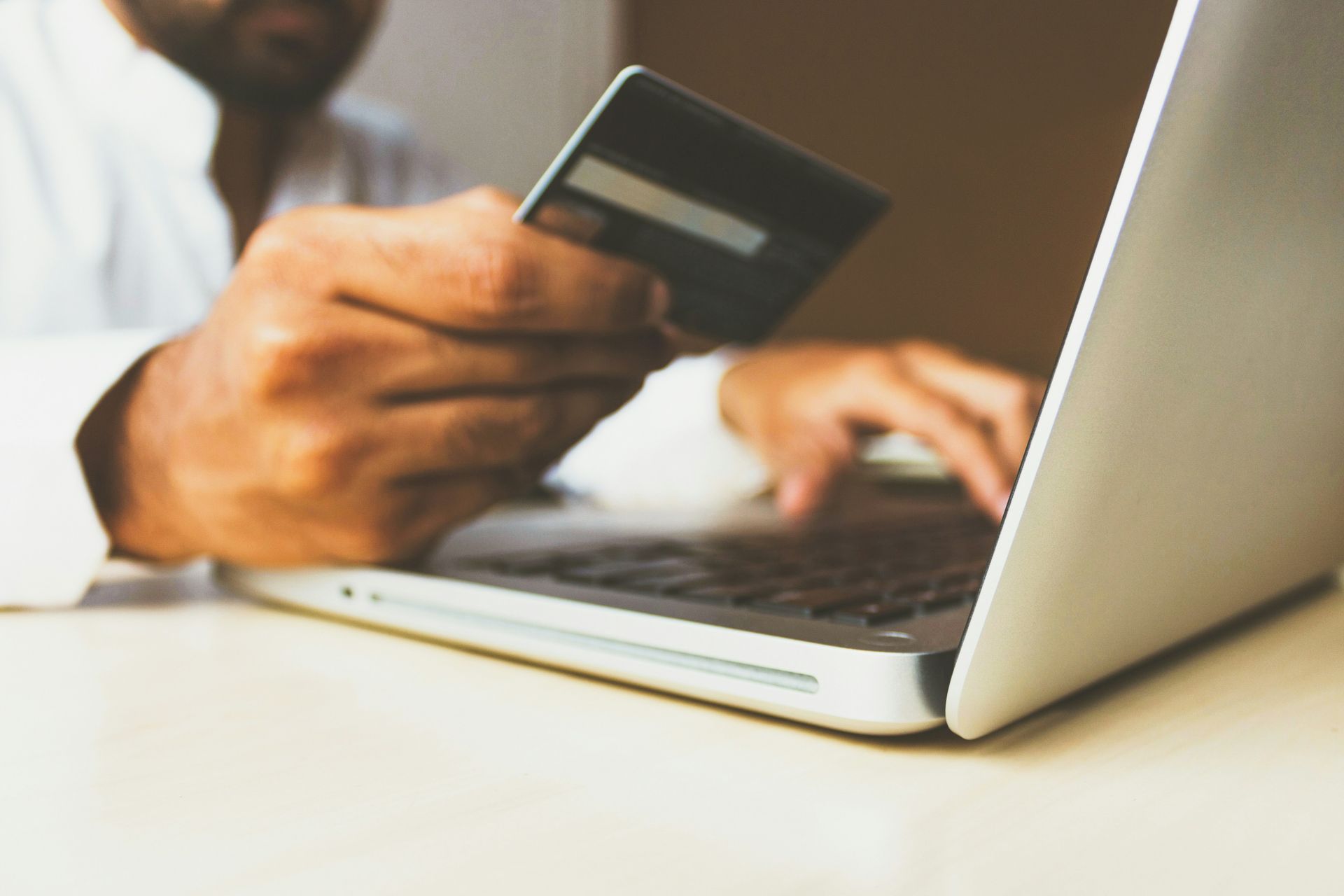 A person in a white shirt sits at a laptop, holding a credit card in one hand while using the keyboard with the other.