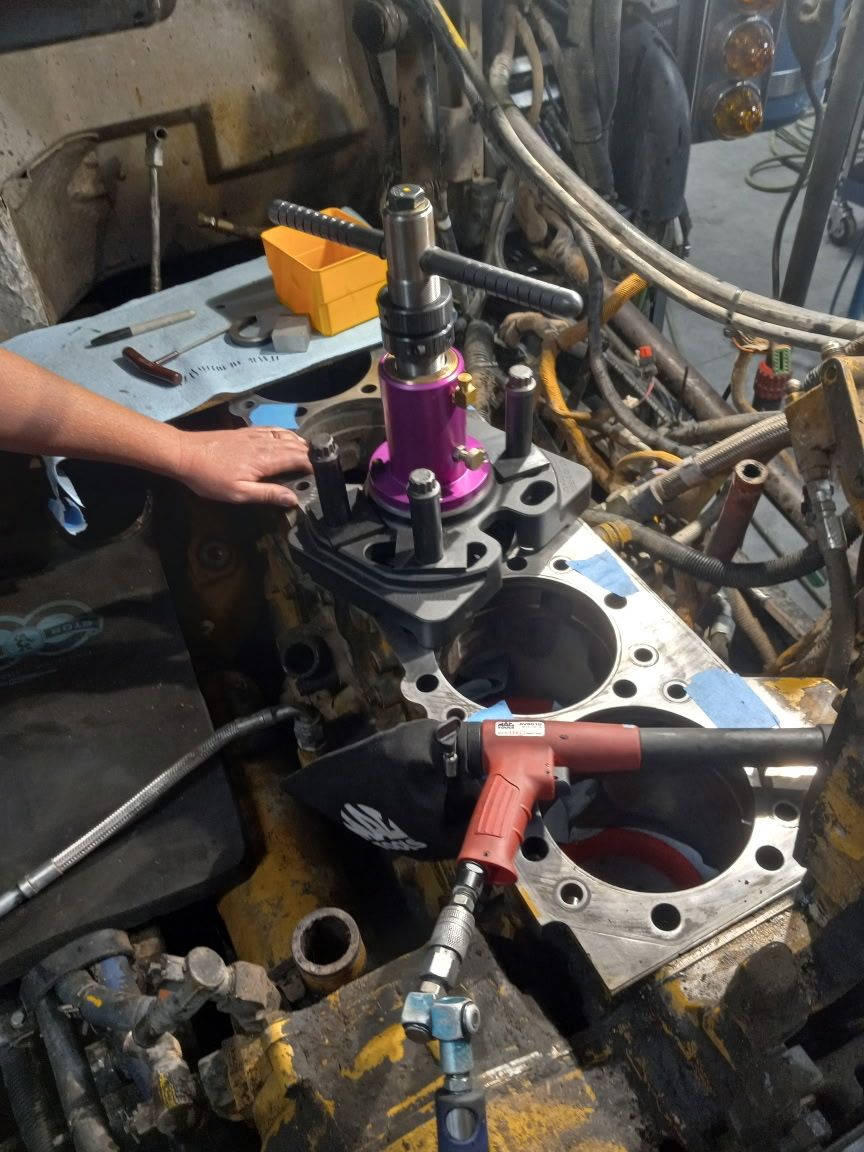 Mechanic kneeling, working on truck engine with hood up. Wearing gloves, overalls, and a cap.