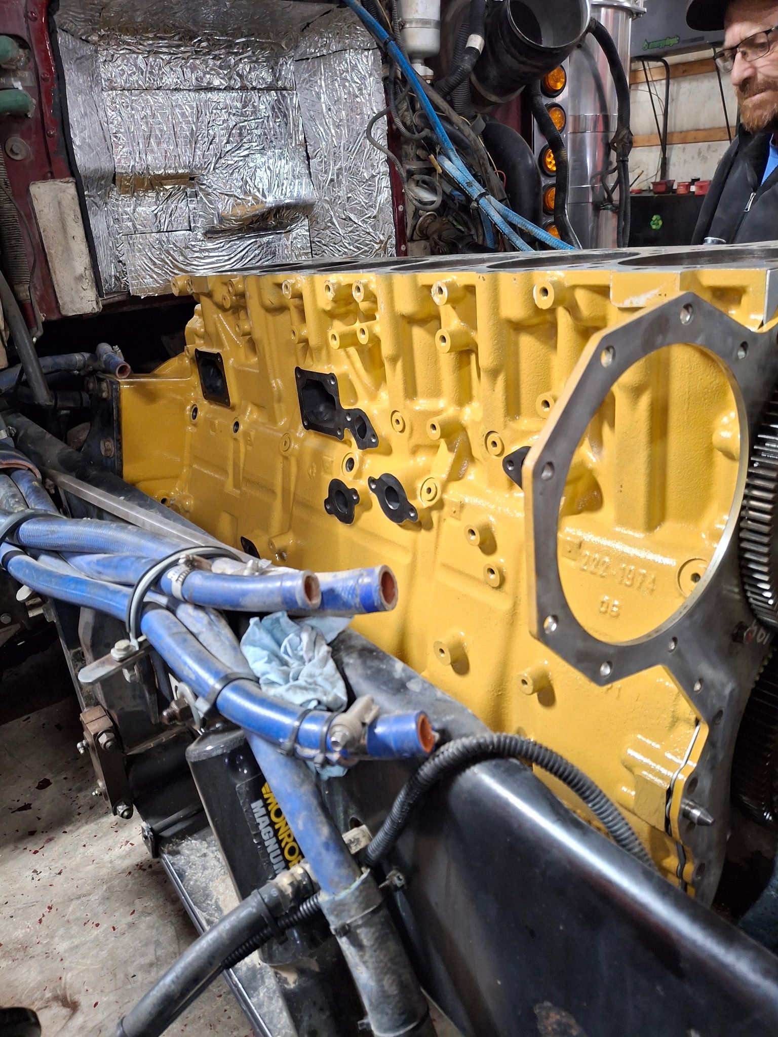 Mechanic kneeling, working on truck engine with hood up. Wearing gloves, overalls, and a cap.