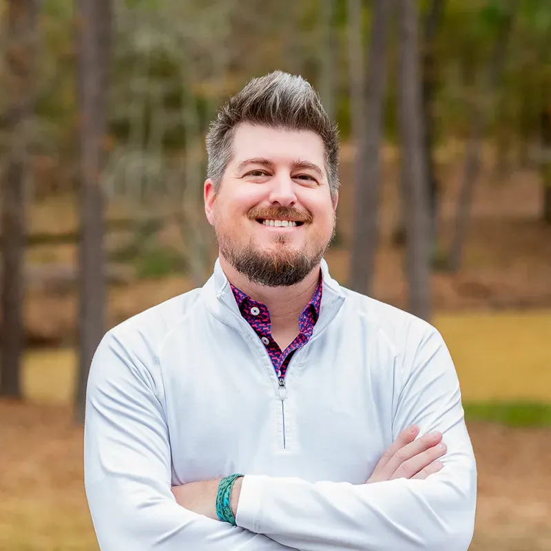 Man with arms crossed, smiling, wearing white zip-up over a patterned shirt, outdoors.
