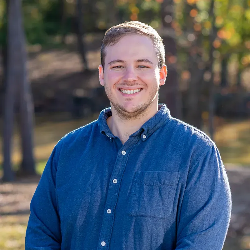Smiling man in blue button-down shirt outdoors, blurred trees in background.