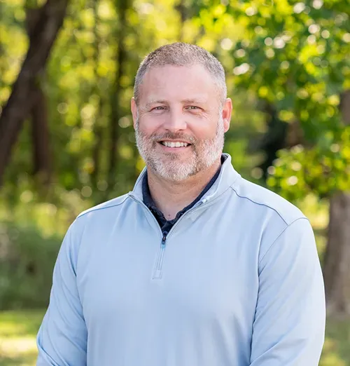 Man with graying beard smiles in light blue sweater, outdoor setting with trees.