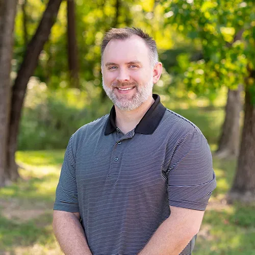 Man with a trimmed beard smiling outdoors, wearing a gray striped polo shirt; trees in the background.