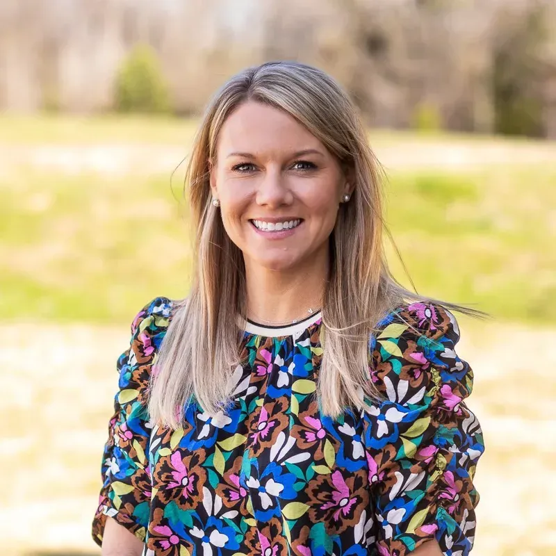 Woman smiling, wearing colorful floral blouse, standing outside.