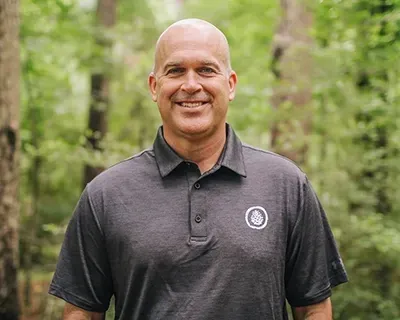 A bald man wearing a black polo shirt is standing in the woods.