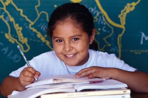 A young girl is smiling while sitting at a desk with books and a pen