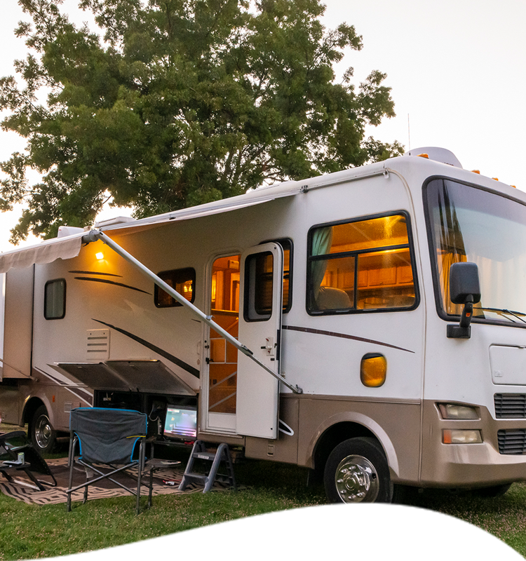 RV parked outdoors with awning extended, lights on inside, and a chair set up nearby.