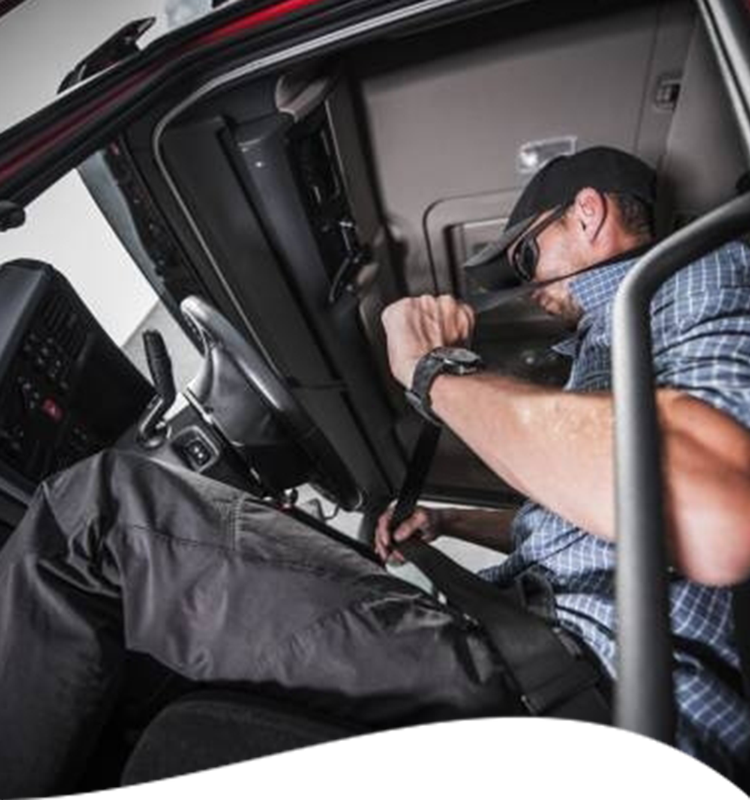 A truck driver fastens his seatbelt in the cab. He wears a cap, glasses, and a plaid shirt.