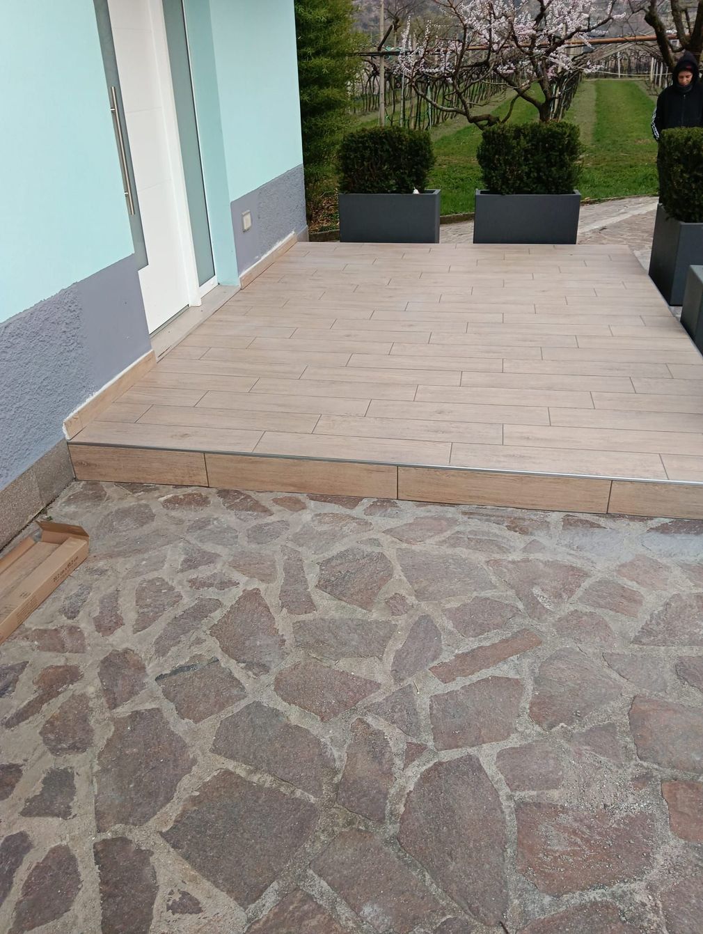 Patio in pietra che conduce a una terrazza in legno chiaro adiacente a un edificio, con fioriere e siepi nere sullo sfondo.
