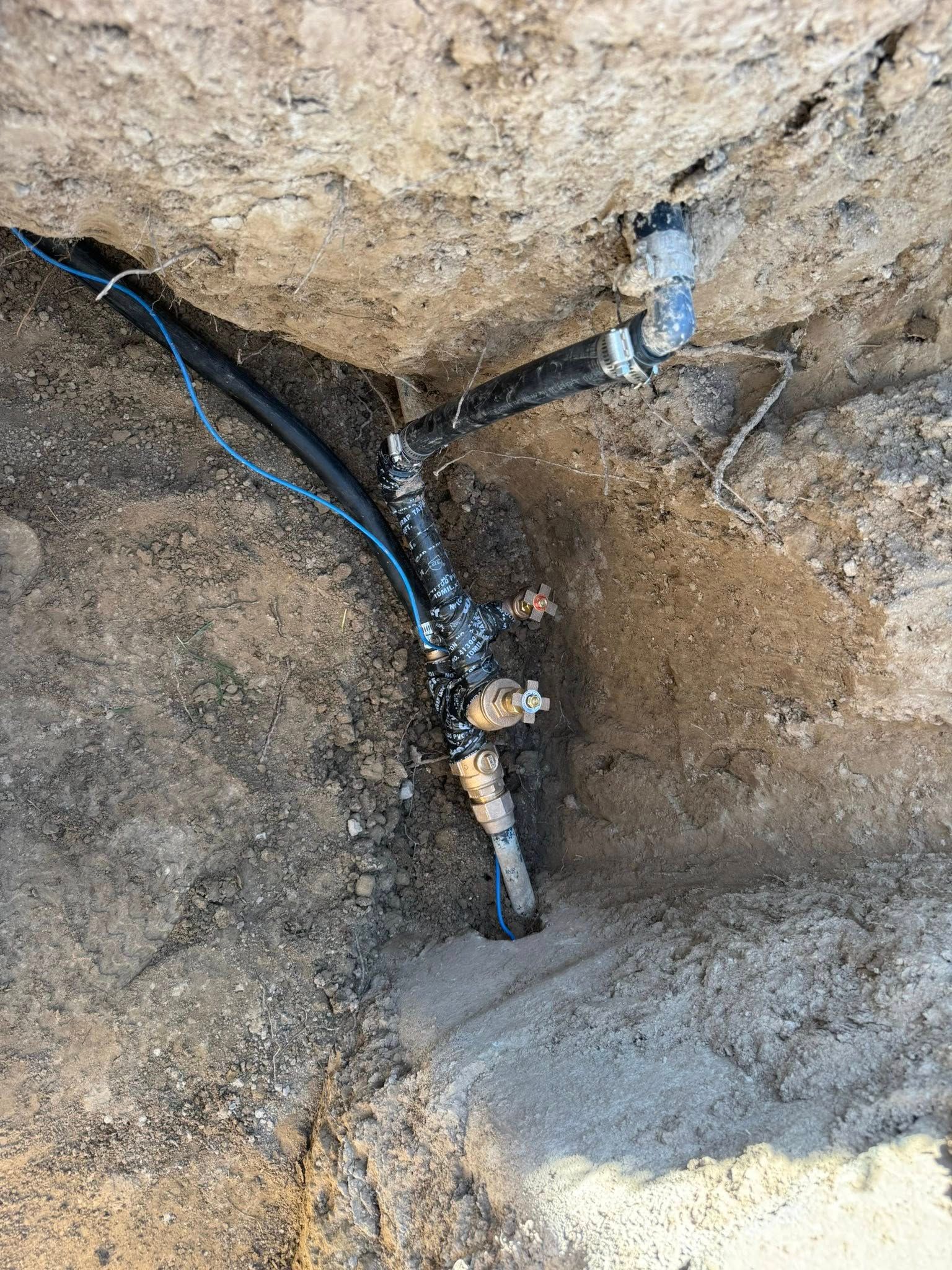 Exposed electrical wires and a damaged pipe in a dirt trench against a rock wall