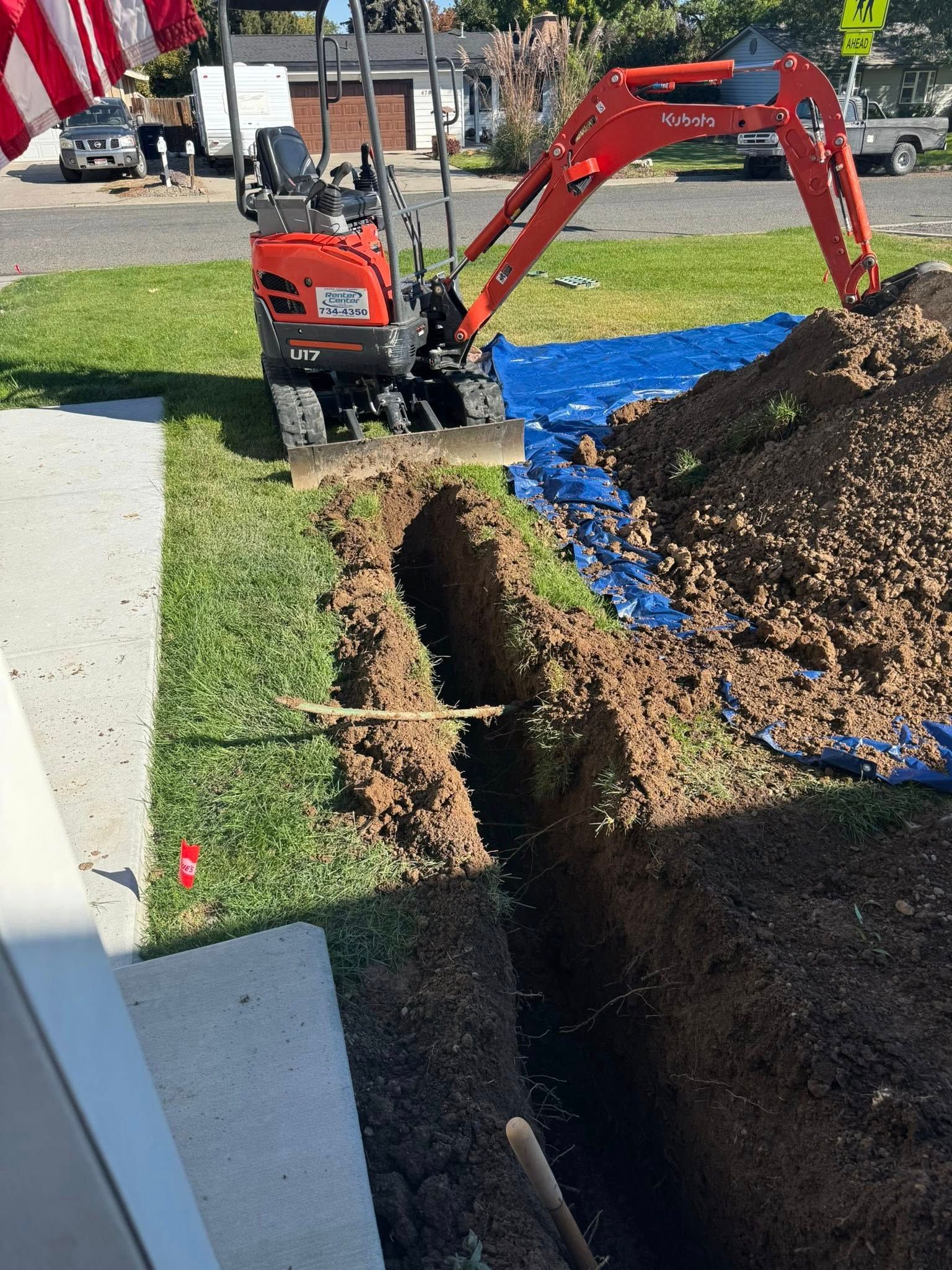Red mini excavator digging a trench beside a sidewalk with a blue tarp and dirt pile nearby.