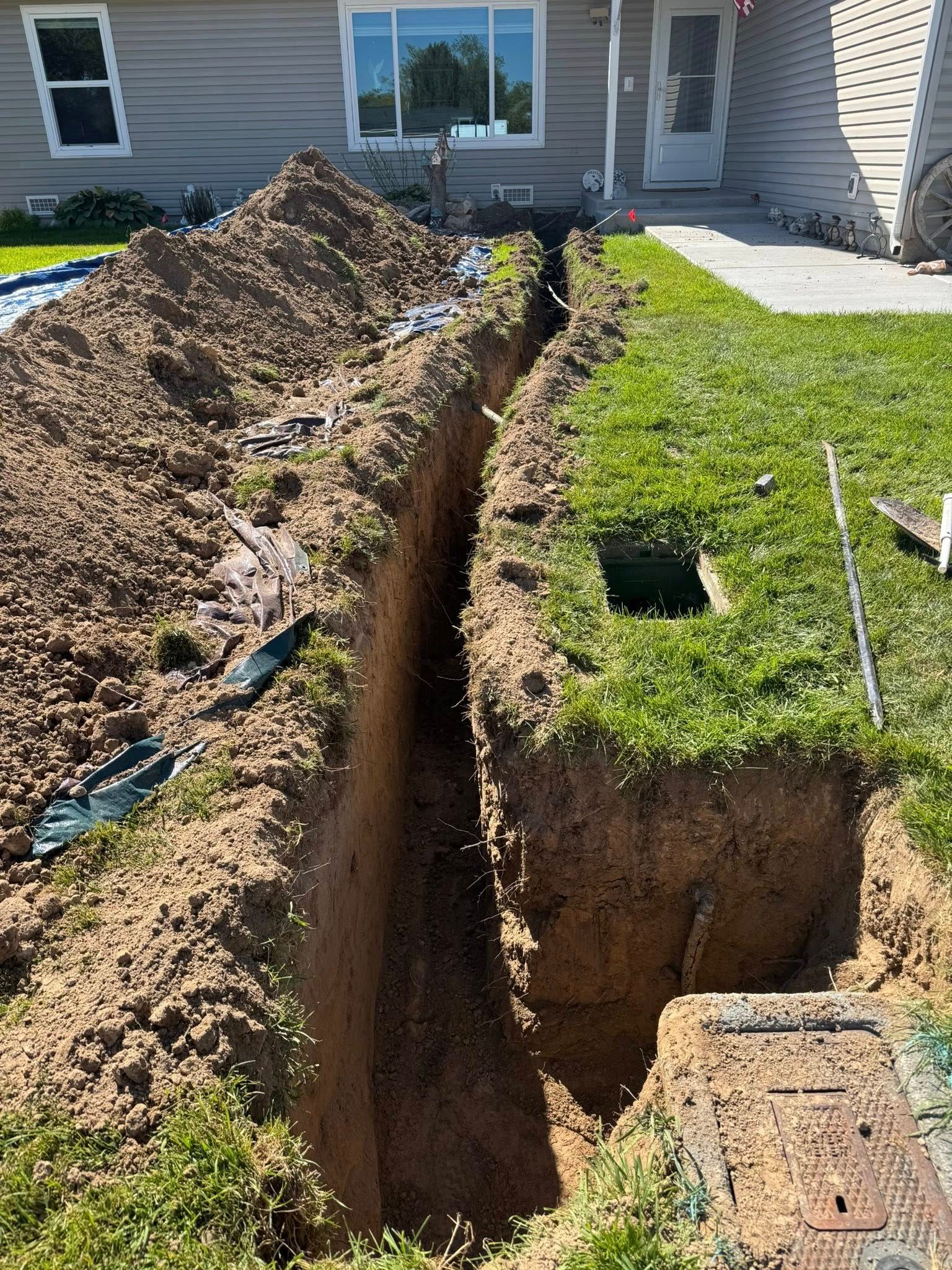 Backyard trench beside a house, with piles of excavated dirt and exposed utility holes in the grass.