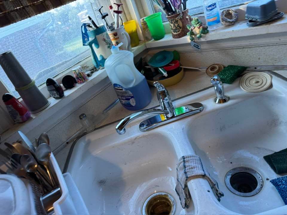 A cluttered kitchen sink with dishes, dish soap, and cleaning items on the counter by a window.