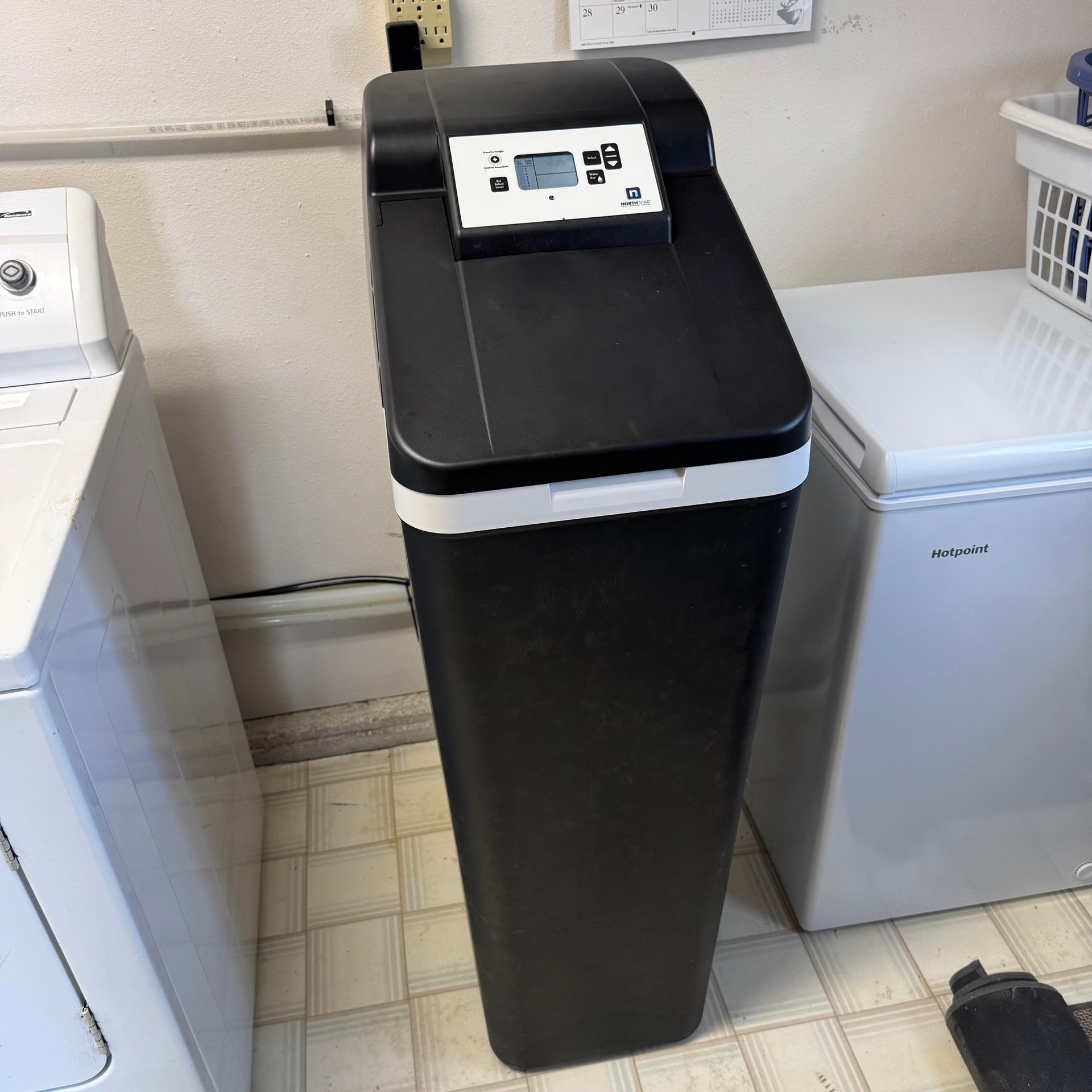 Black trash bin with digital control panel between a washing machine and chest freezer in a laundry room