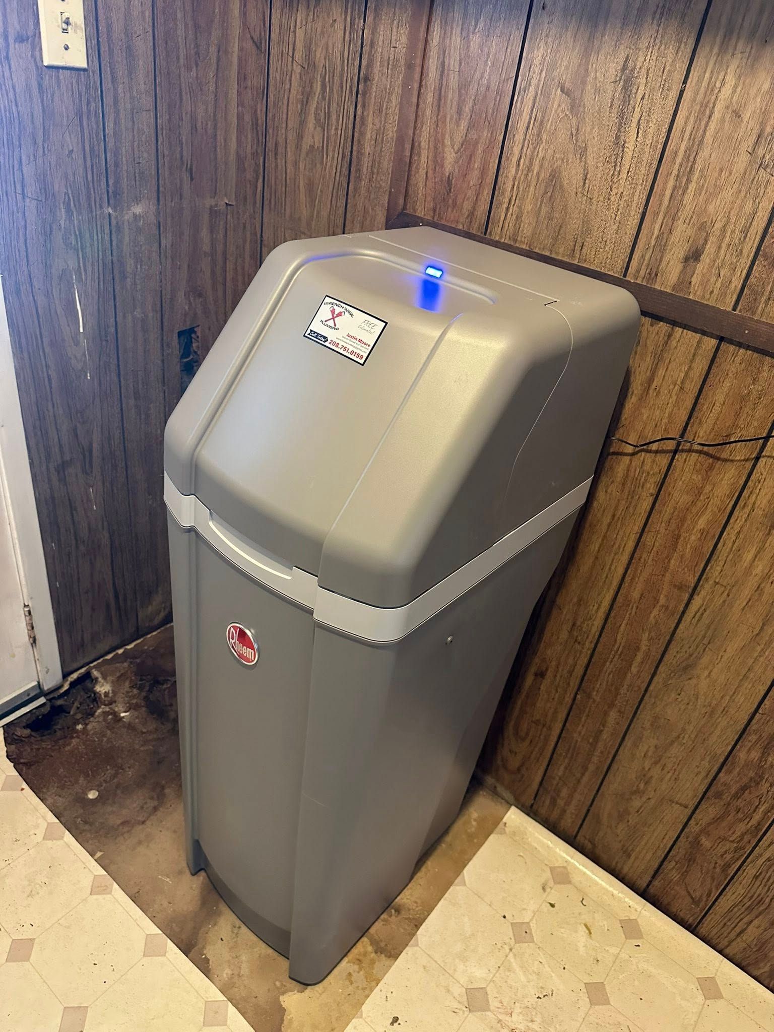 Gray trash compactor in a wood-paneled corner, with a blue light on top.