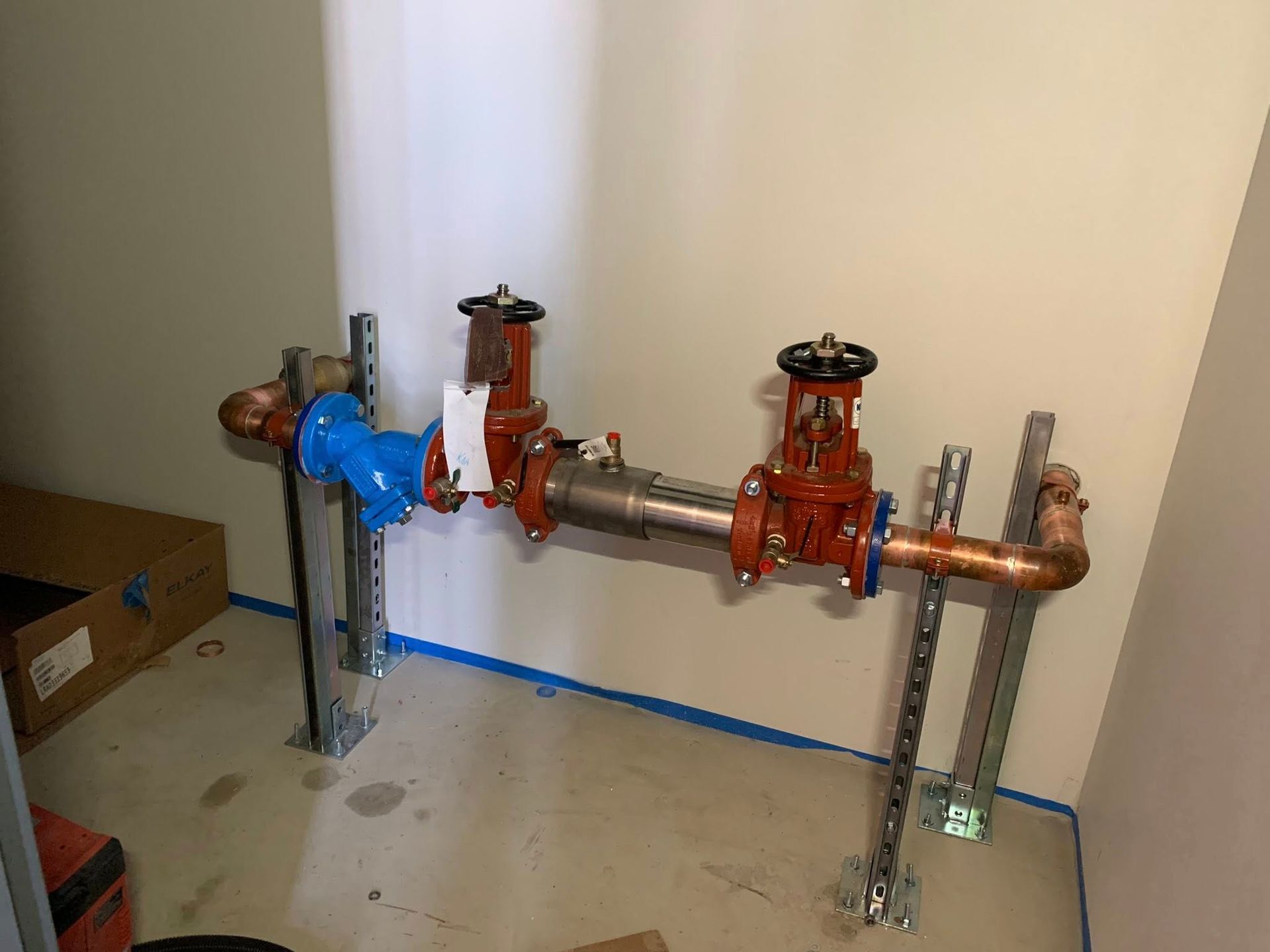 Industrial piping and valves against a wall, with a blue valve assembly and copper pipes on a concrete floor
