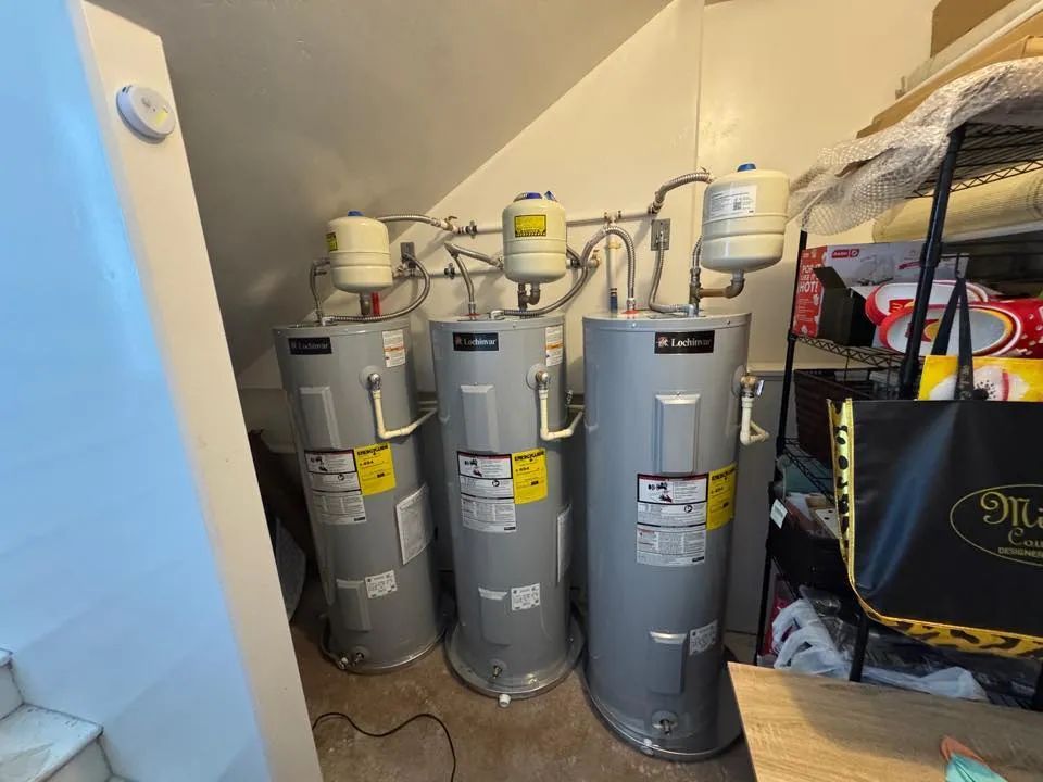 Three gray water heaters and plumbing pipes in a small utility closet