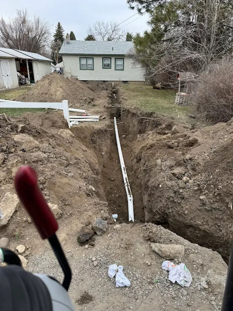 Deep trench dug across a yard toward a small house, with exposed white pipe and dirt piles.