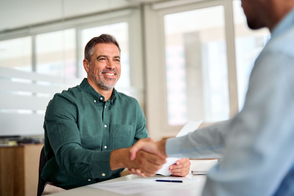 Man in green shirt smiles, shaking hands with another person at a desk in an office.