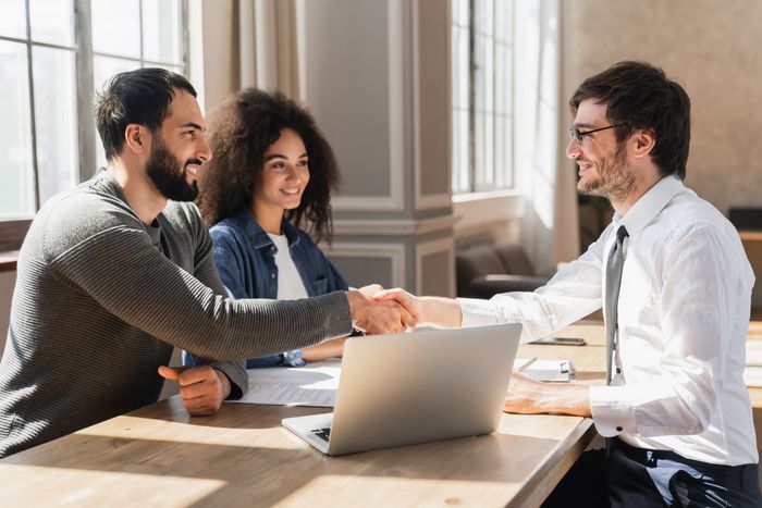 A couple shakes hands with a professional over a table with a laptop, smiling.