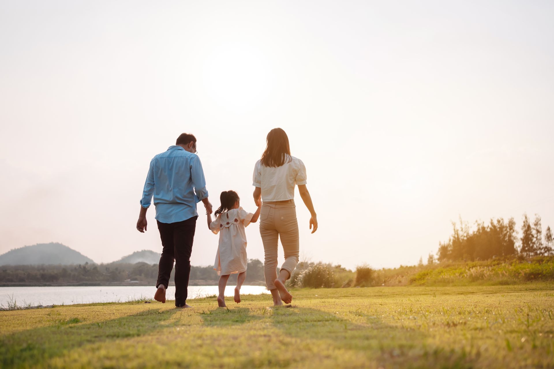 Family walking away from the camera, holding hands, along a grassy bank near water. Sunlight.