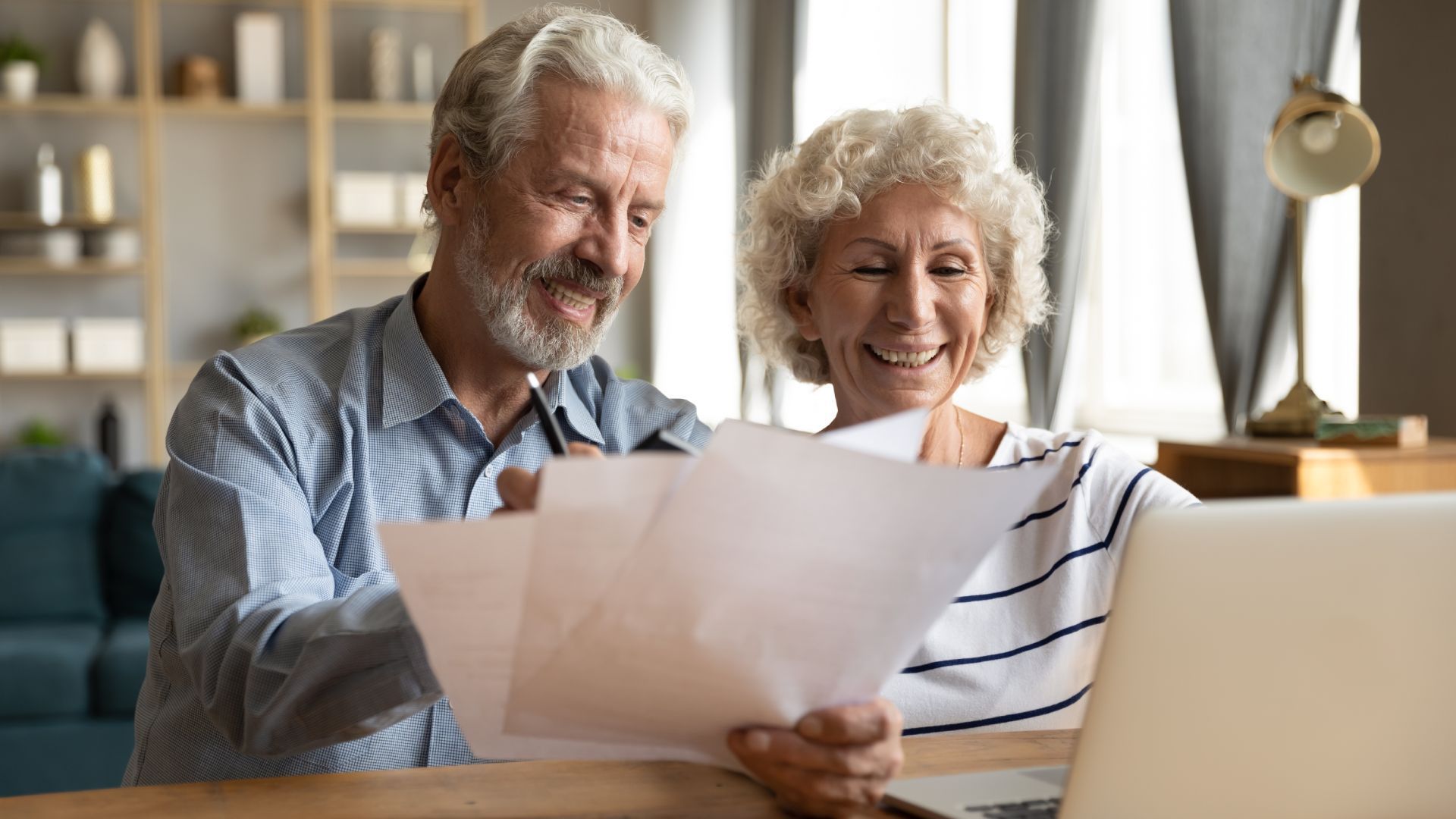 Couple reviewing papers together, smiling; interior, laptop visible.