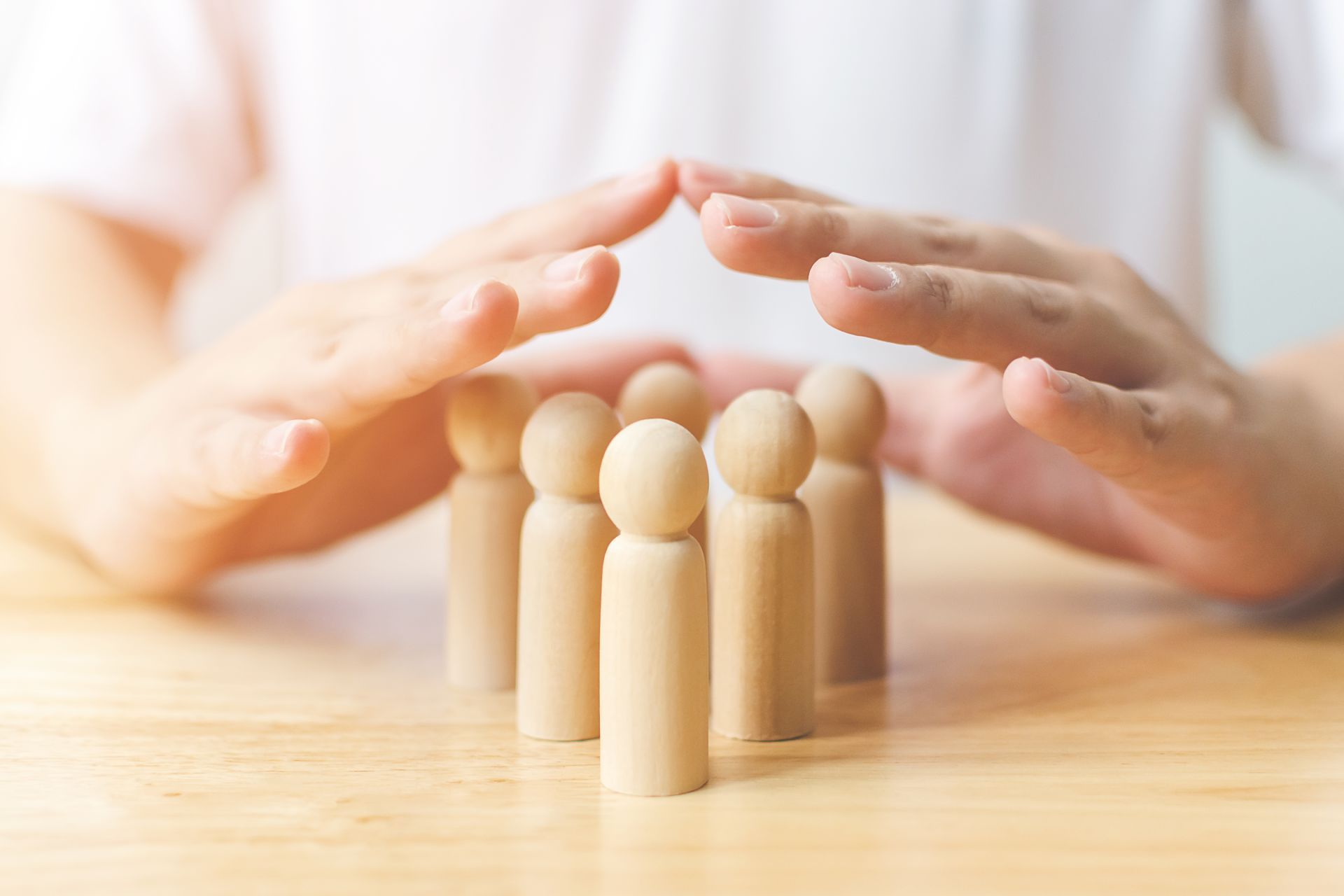 Hands sheltering wooden figures on a wooden surface, symbolizing protection and care.
