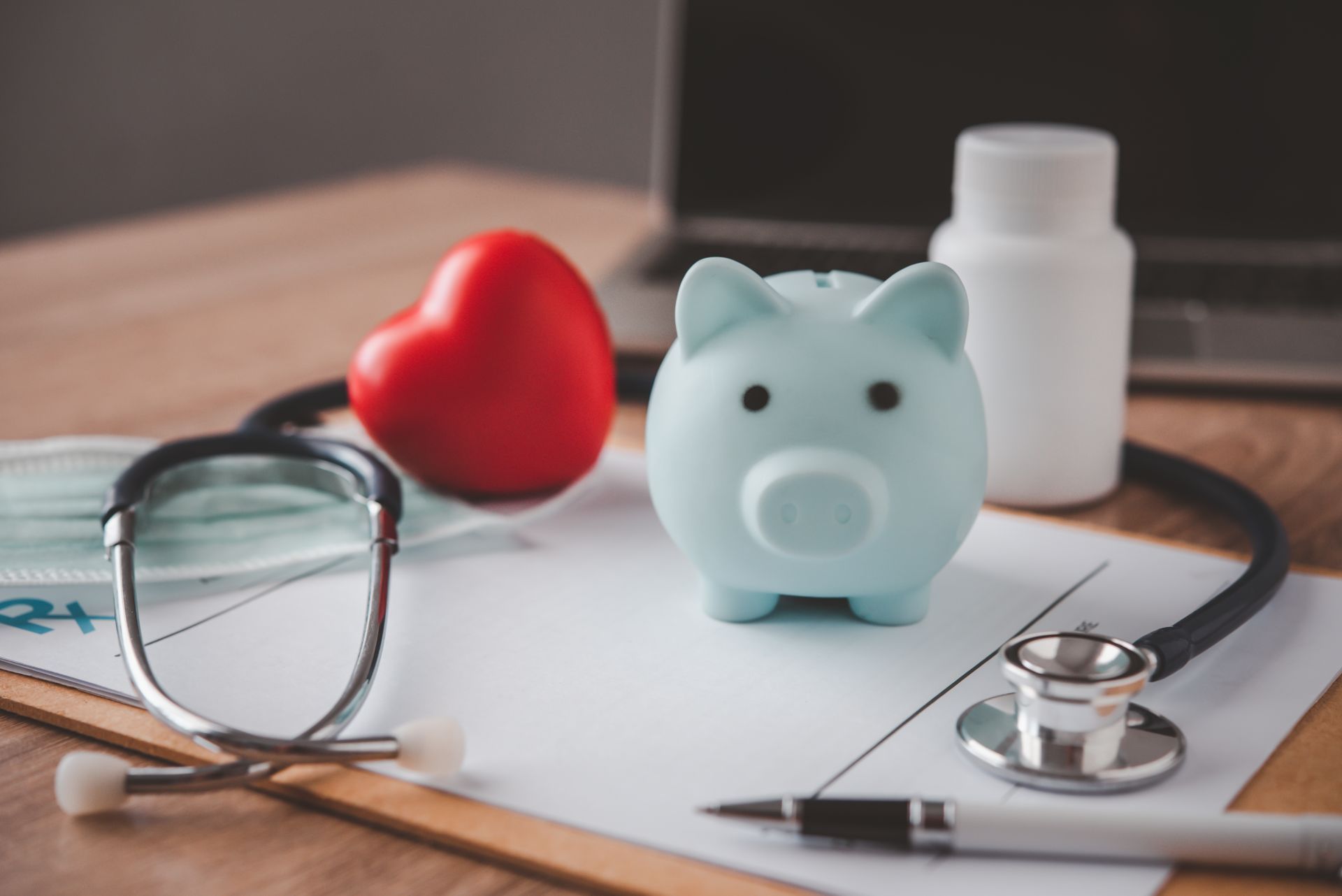 Piggy bank, stethoscope, red heart, and prescription on a desk, symbolizing healthcare costs.