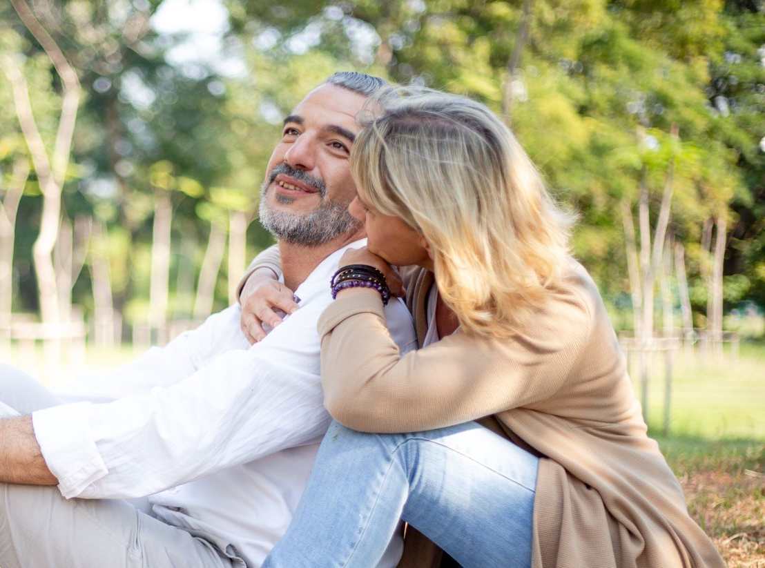 Couple sitting outdoors, embracing. Man looks upward, smiling. Woman leans on him. Green trees background.