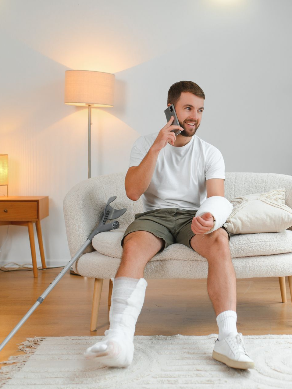 Man with arm and leg casts sits on sofa, talking on phone, crutch nearby.