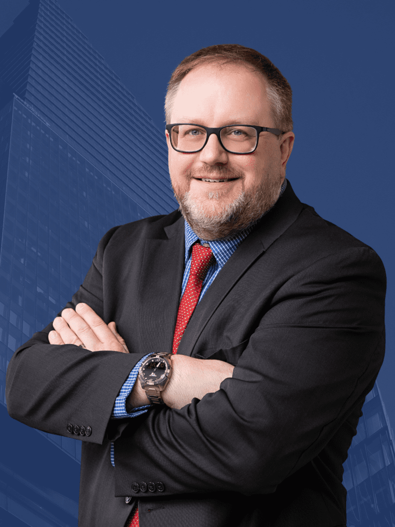 Man in a suit with arms crossed, smiling. Red tie, glasses. Blue background with skyscrapers.