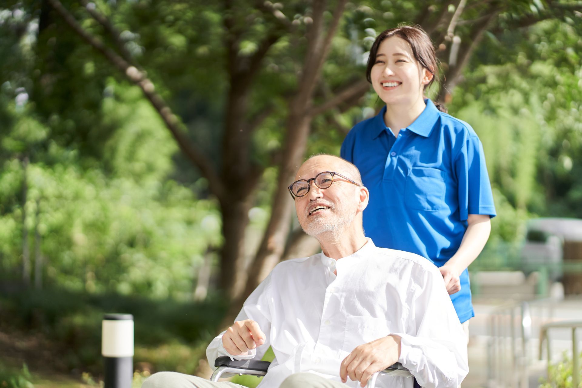 A caregiver in a blue polo shirt walks behind an individual in a wheelchair through a park, both looking up and smiling.