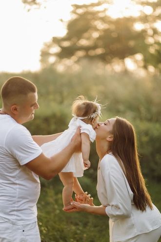 A parent holding a toddler while a second person leans in to kiss the child in a sunlit field.